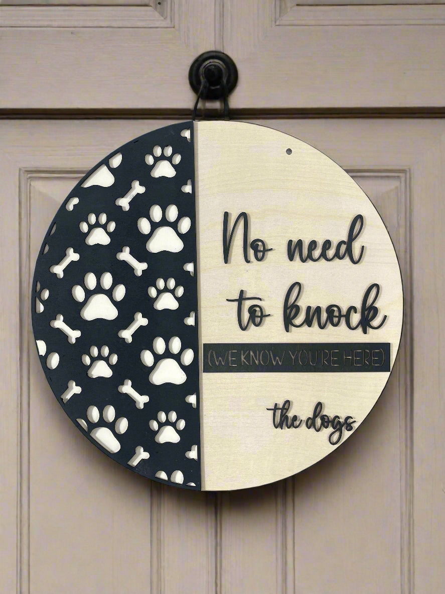 A round wooden sign with black and white design featuring paw prints, bones, and text that reads 'No need to knock - the dogs'.